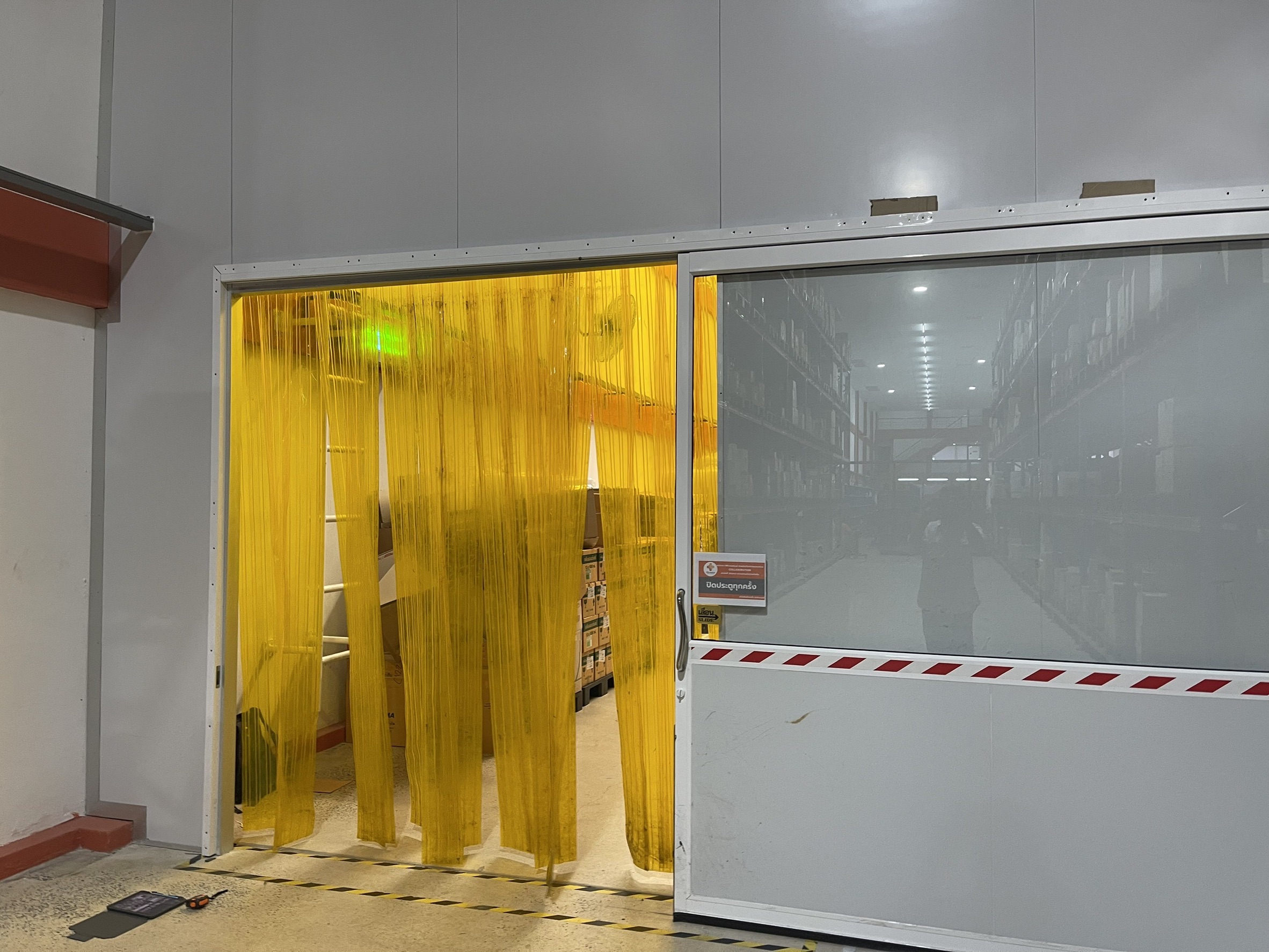 ม่านริ้ว PVC สีเหลืองแขวนในคลังสินค้า เคียงข้างประตูม้วน PVC XEKA ผลงานติดตั้งจริง